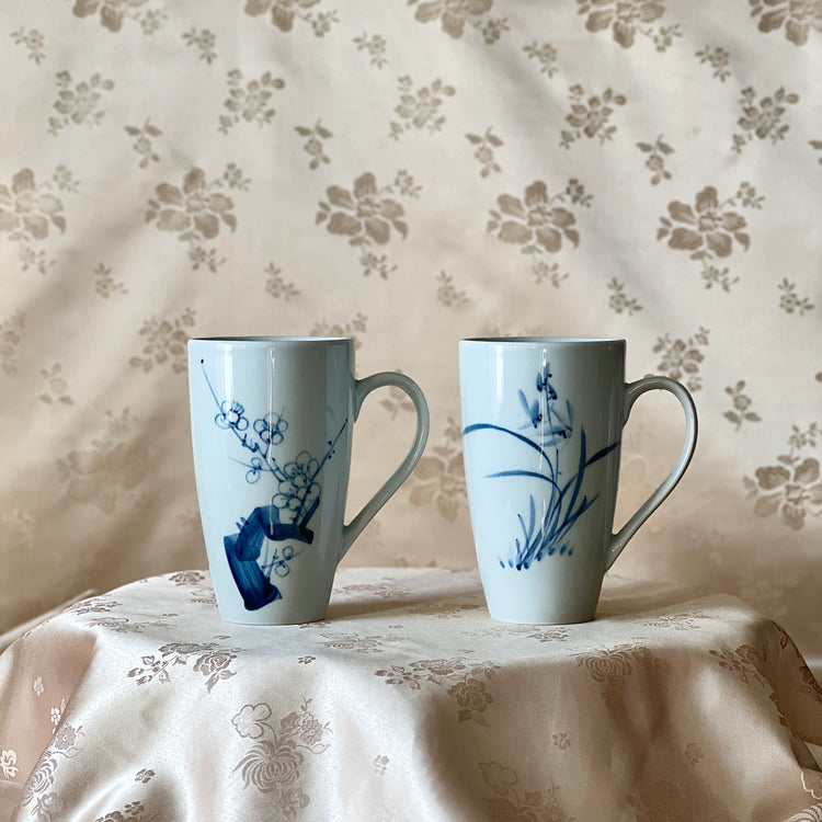 Korean white porcelain mug set with blue plum blossom and orchid motifs displayed on soft floral fabric