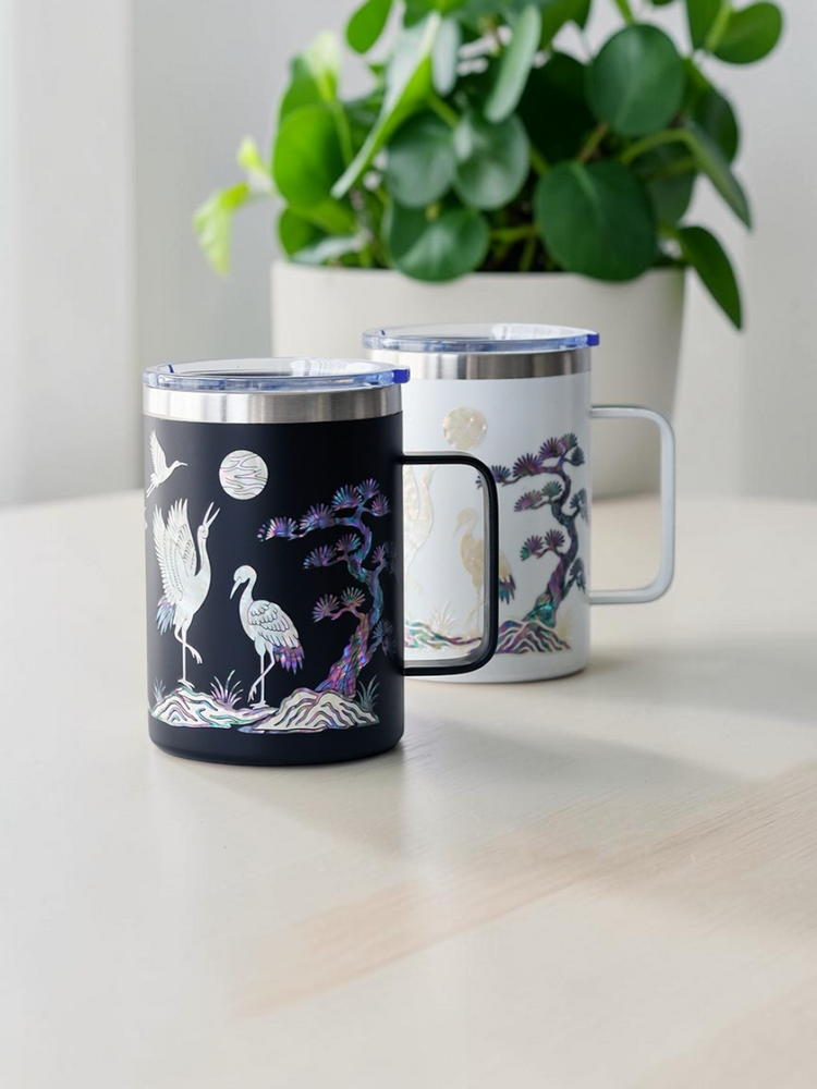 Korean mother of pearl mugs in black and white with crane and pine inlay, styled beside a green houseplant