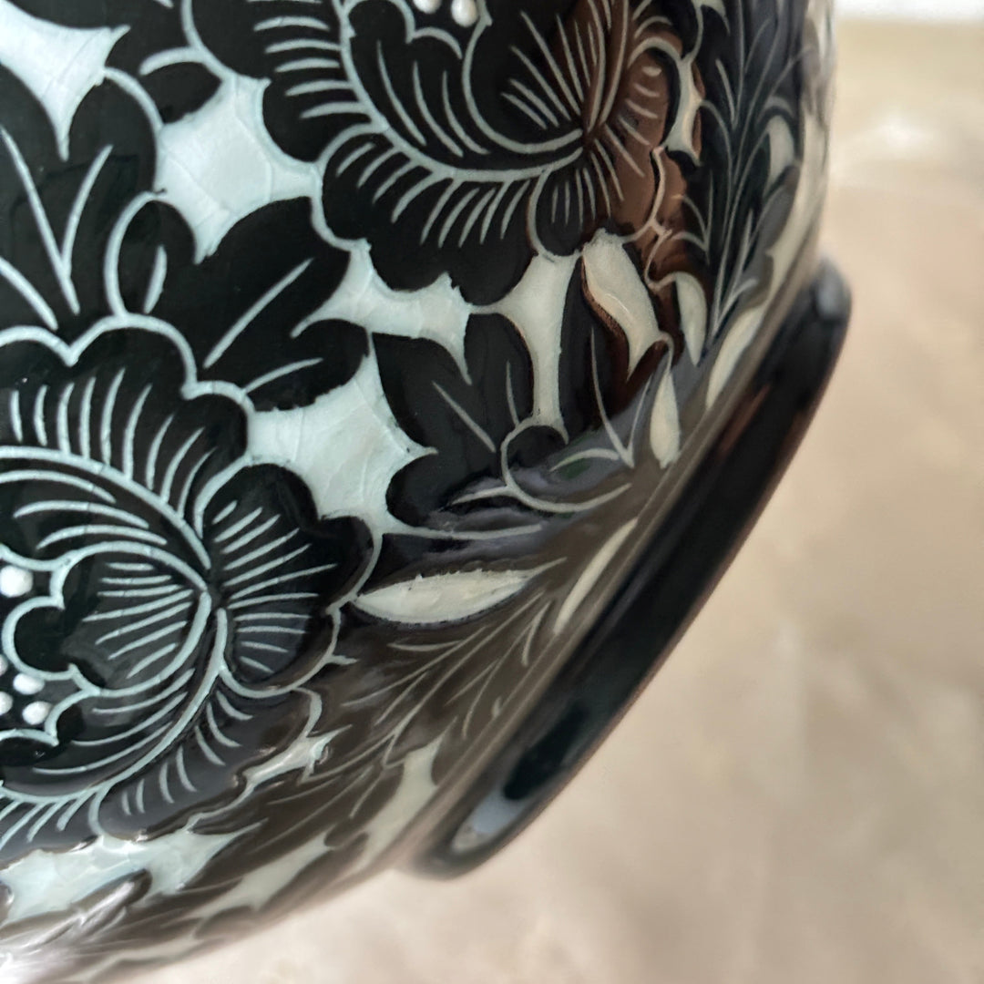 Close-up of a glossy ceramic vase or bowl featuring a black floral pattern over a white glaze, with a dark base, photographed against a light beige background.