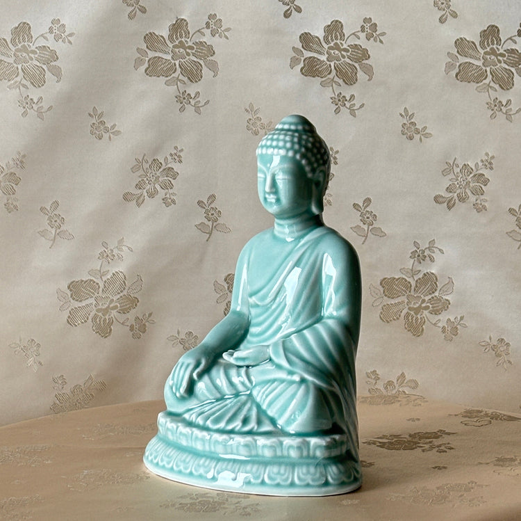 A small seated Buddha figurine in glossy light celadon ceramic, placed on a round table against a beige floral-patterned fabric backdrop.