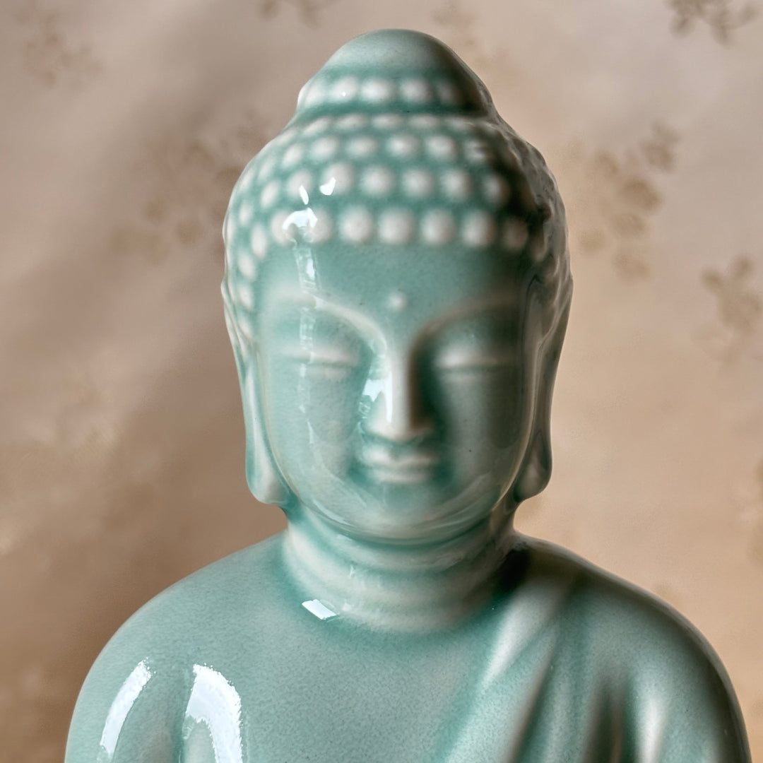 A glossy light blue-green ceramic Buddha statue shown from the chest up against a soft beige patterned backdrop.