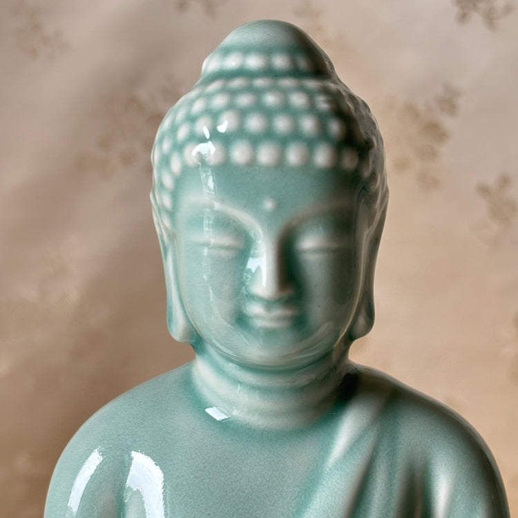 A glossy light blue-green ceramic Buddha statue shown from the chest up against a soft beige patterned backdrop.