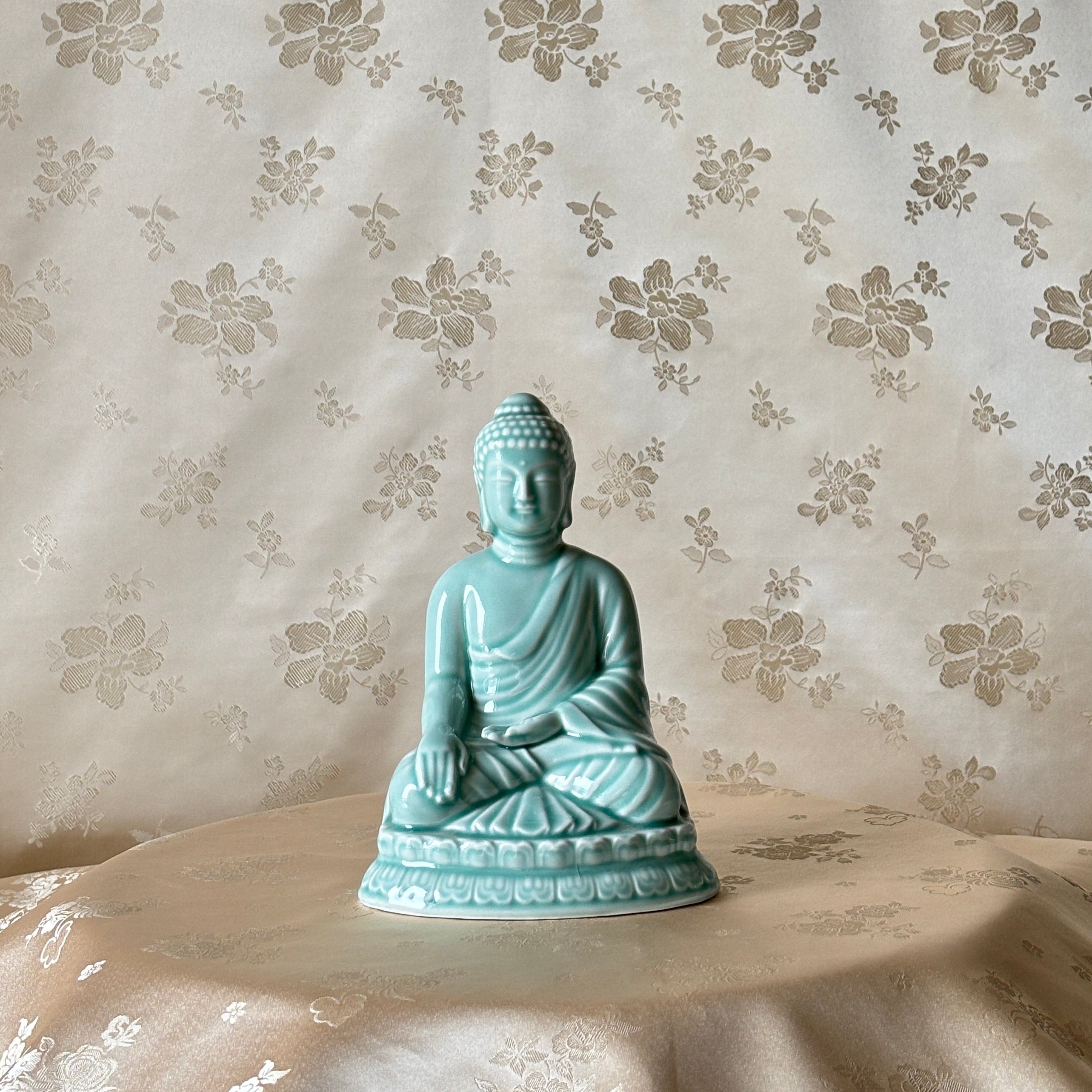 A small seated Buddha figurine in a light aqua or turquoise glazed ceramic finish, displayed on a draped beige fabric surface against a matching beige floral-patterned backdrop.