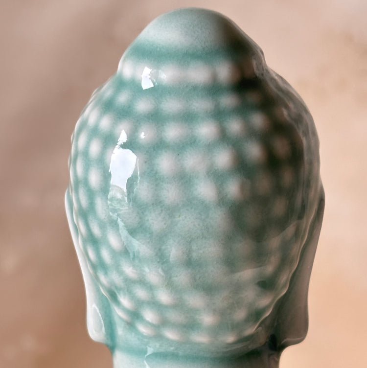 Close-up of the back of a glossy ceramic Buddha bust in a pale aqua or mint green glaze, showing the textured hair pattern and smooth neck against a soft beige background.