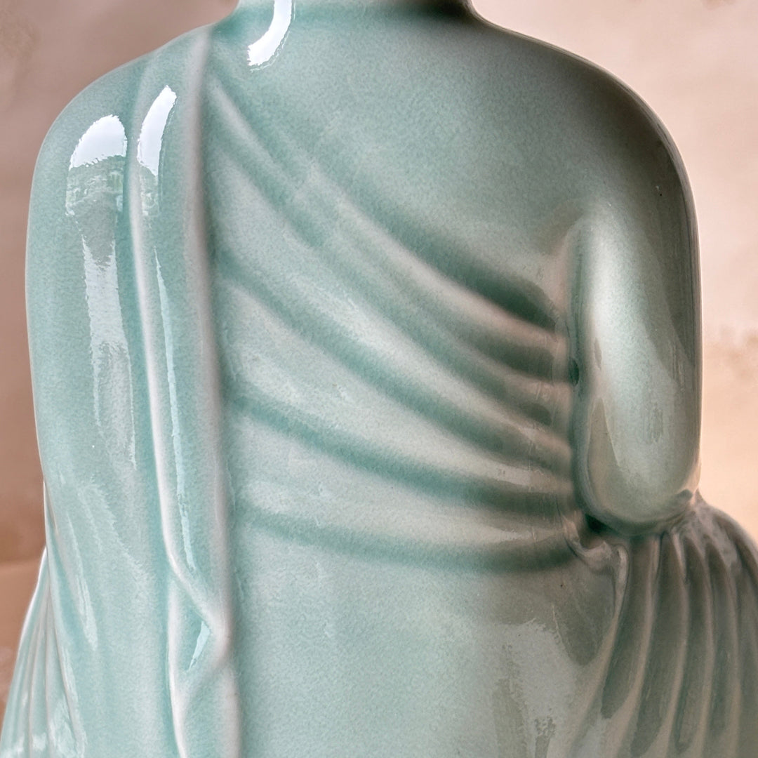 Close-up of a glossy ceramic figurine with a pale aqua-green glaze, showing draped fabric folds and a seated figure from the back.