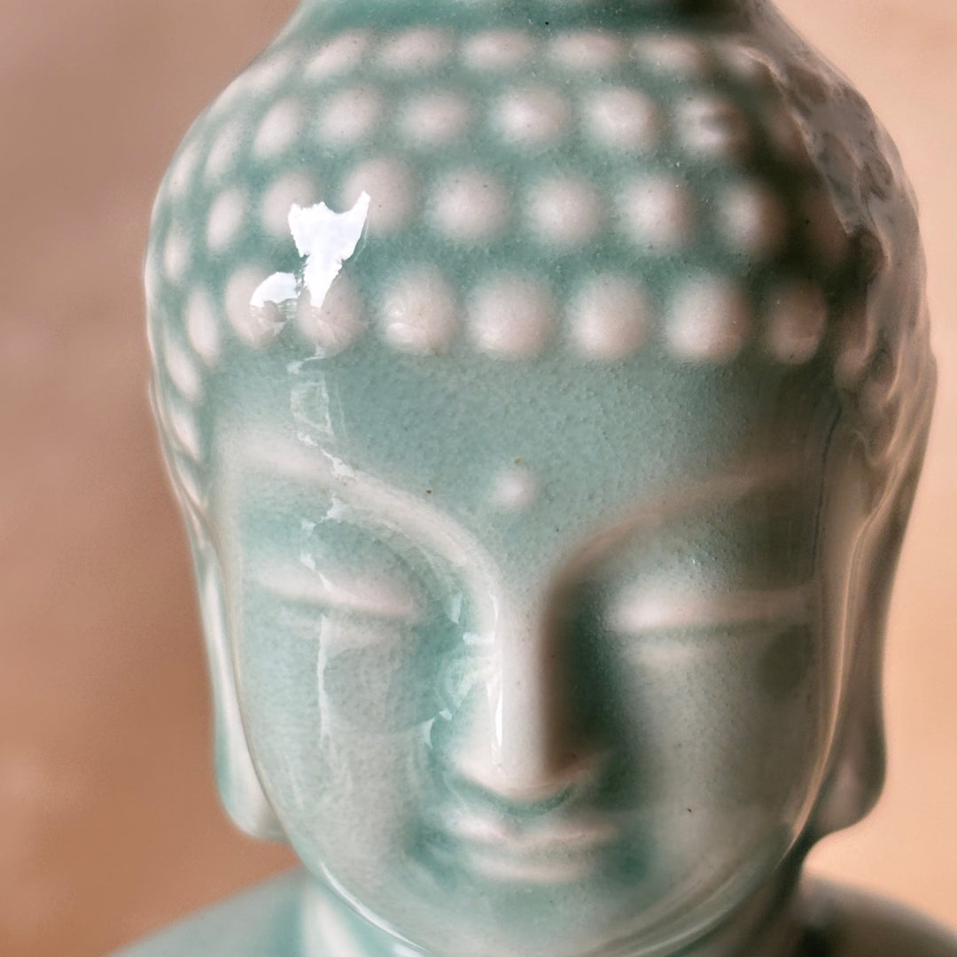 Close-up of a Buddha head statue with a glossy light teal-green glazed finish, showing the detailed ushnisha and serene facial features against a soft warm beige background.