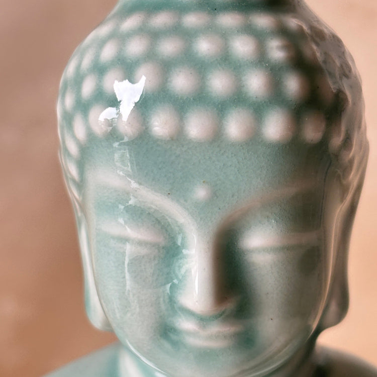 Close-up of a Buddha head statue with a glossy light teal-green glazed finish, showing the detailed ushnisha and serene facial features against a soft warm beige background.