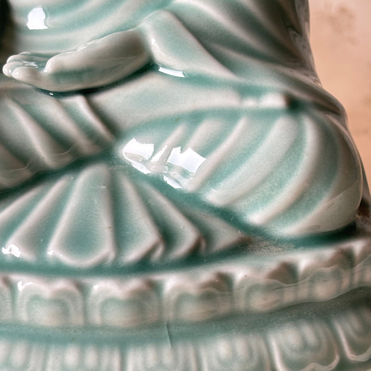 Close-up of a glossy ceramic Buddha figurine in a celadon glaze, showing carved drapery and a decorative base against a soft beige background.