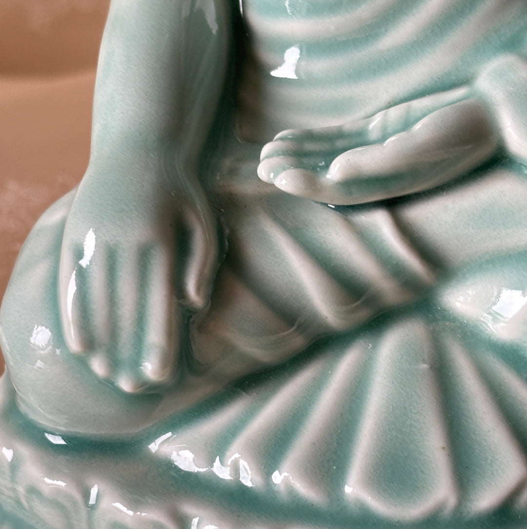 Close-up of a seated ceramic figurine with a glossy pale aqua glaze, showing folded hands and a lotus-style base.