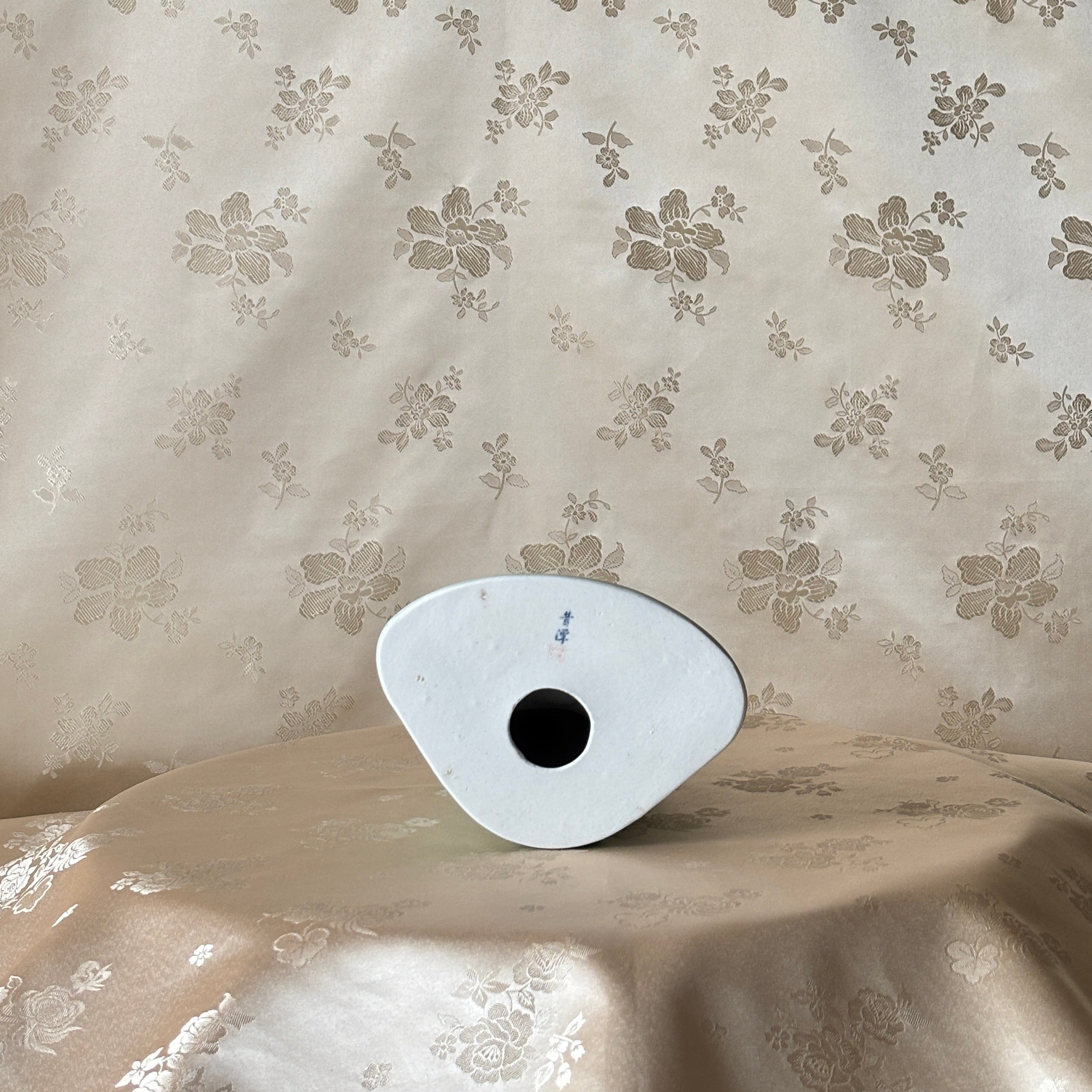 A small white oval object with a black circular center sits on draped beige fabric in front of a matching beige floral patterned backdrop.
