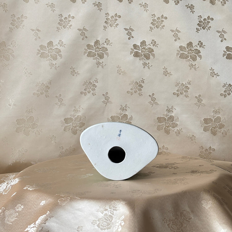 A small white oval object with a black circular center sits on draped beige fabric in front of a matching beige floral patterned backdrop.
