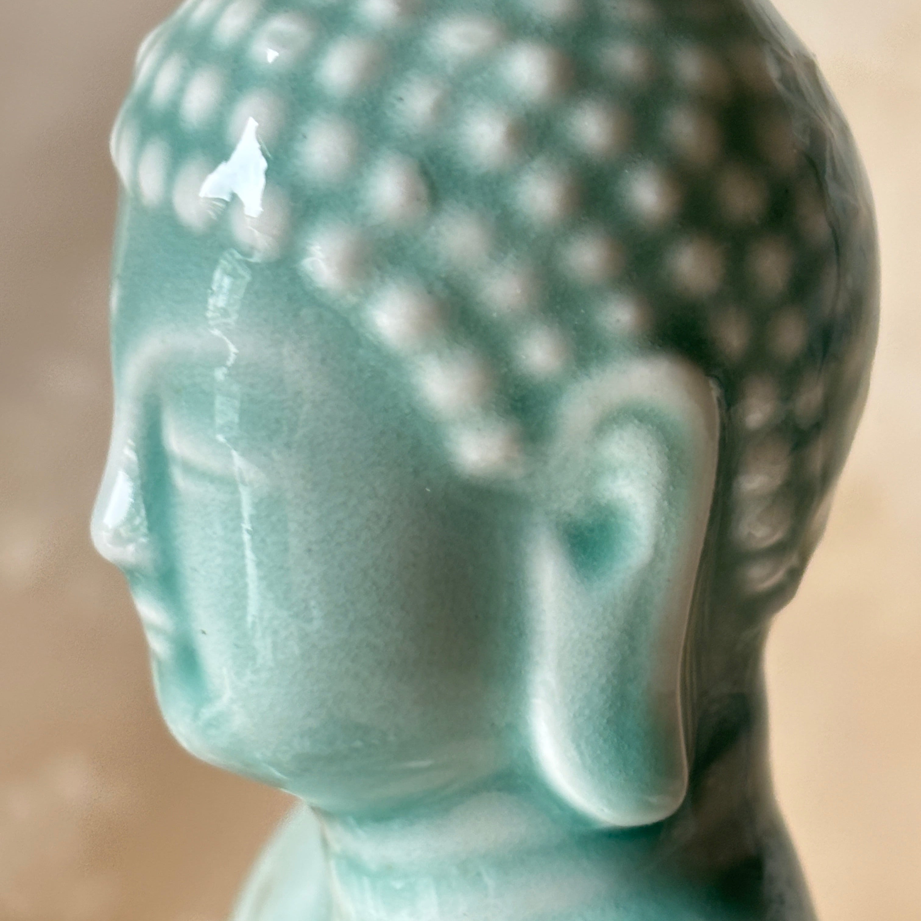 Close-up of a Buddha head statue in a glossy light aqua or turquoise glaze, shown in profile against a soft beige background.