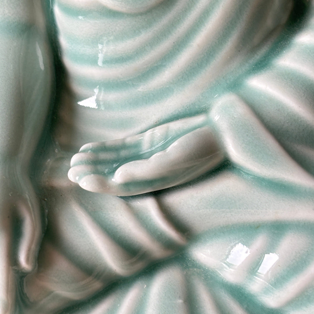 Close-up of a glossy ceramic Buddha statue with a pale aqua glaze, showing detailed folds in the robes and a hand resting in a meditation pose.
