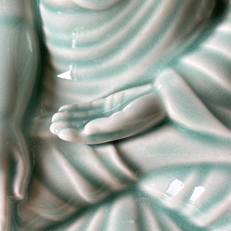 Close-up of a glossy ceramic Buddha statue with a pale aqua glaze, showing detailed folds in the robes and a hand resting in a meditation pose.
