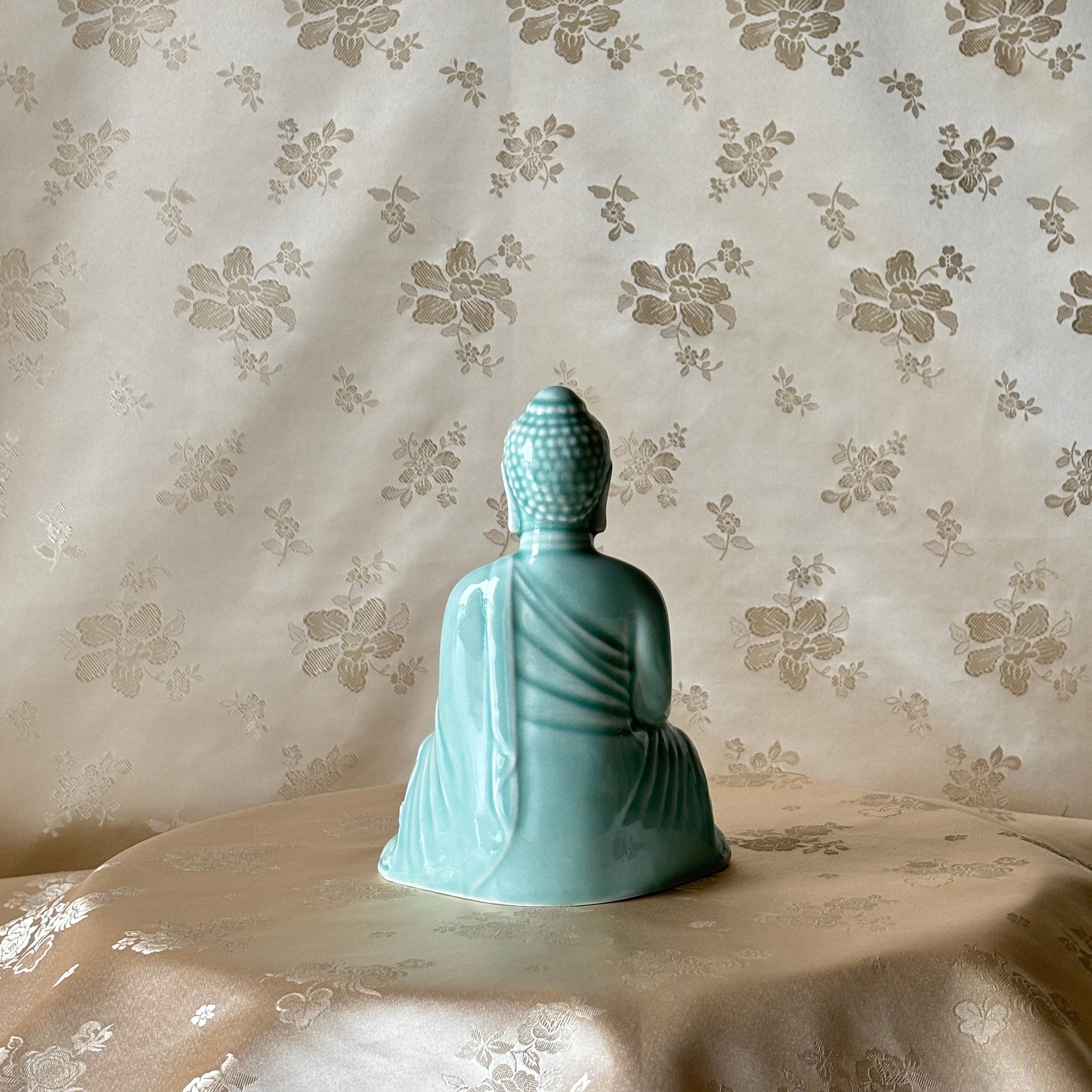 Korean handmade Buddha statue in a light celadon color, shown from the back, sitting on a draped beige satin cloth against a beige floral patterned backdrop.