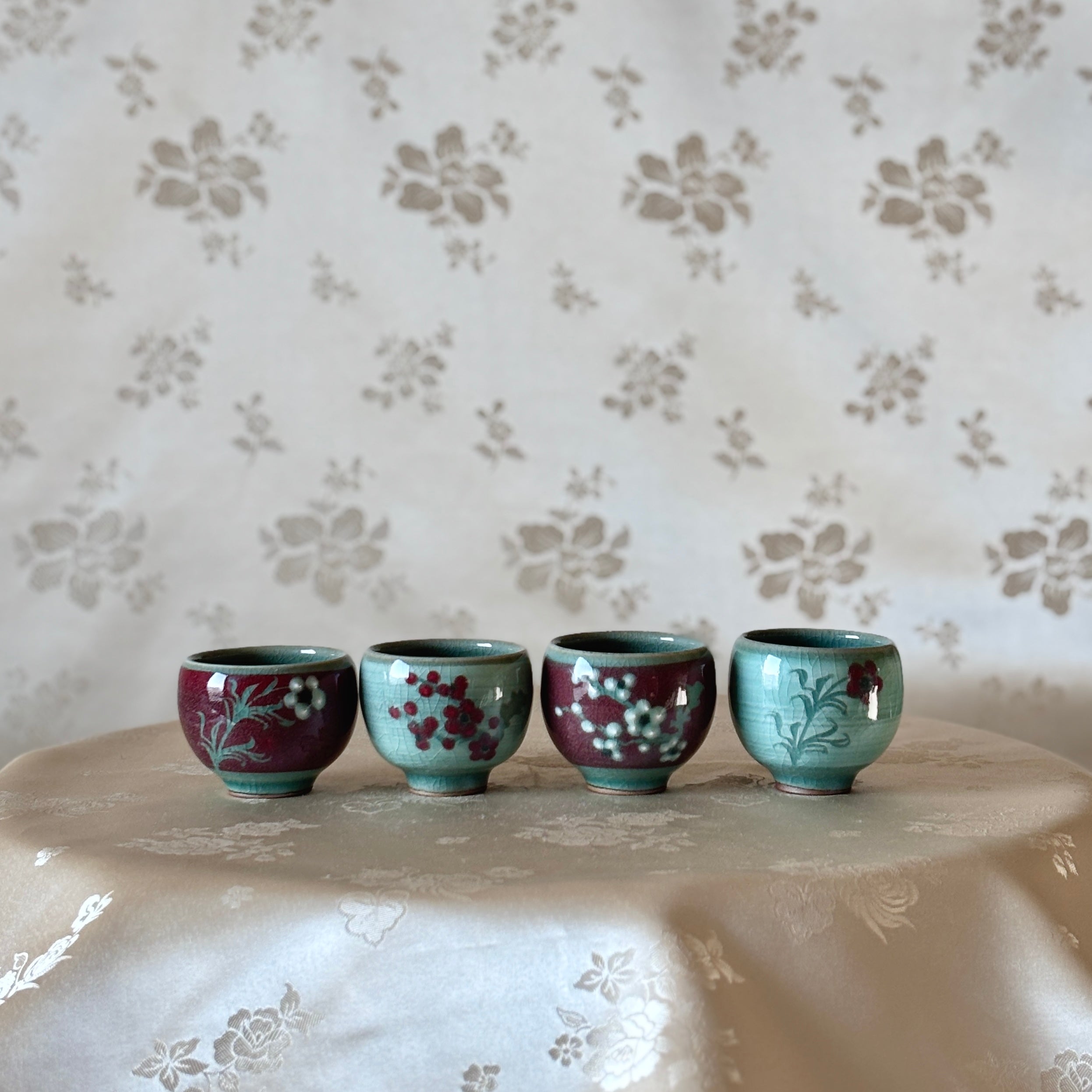 A set of four handcrafted celadon cups with distinct color inlays, displayed on a table with a floral pattern in the background.