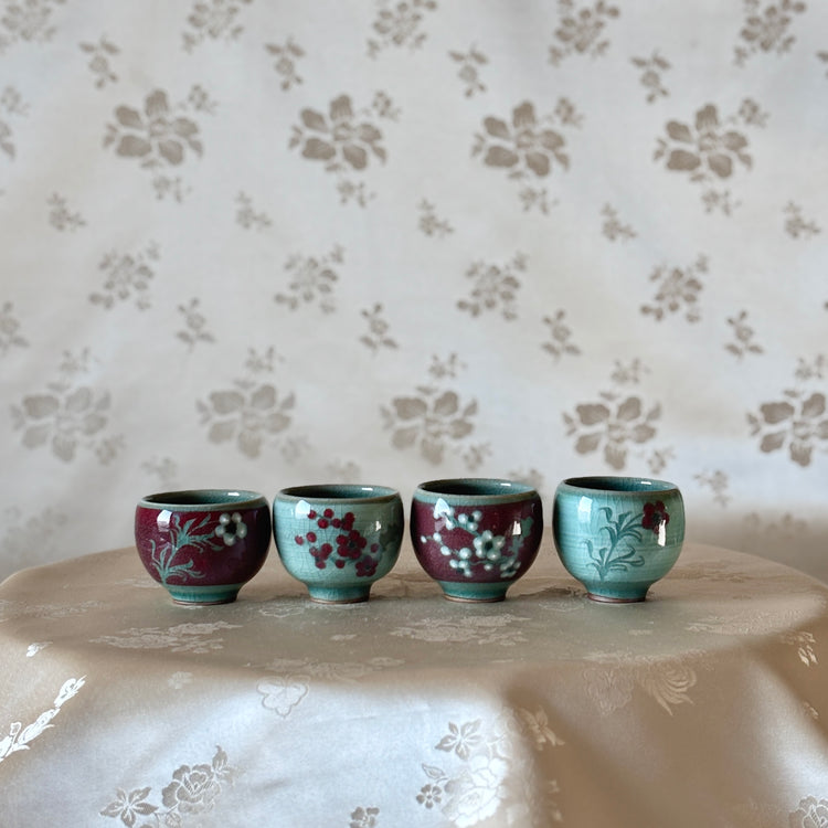 A set of four handcrafted celadon cups with distinct color inlays, displayed on a table with a floral pattern in the background.