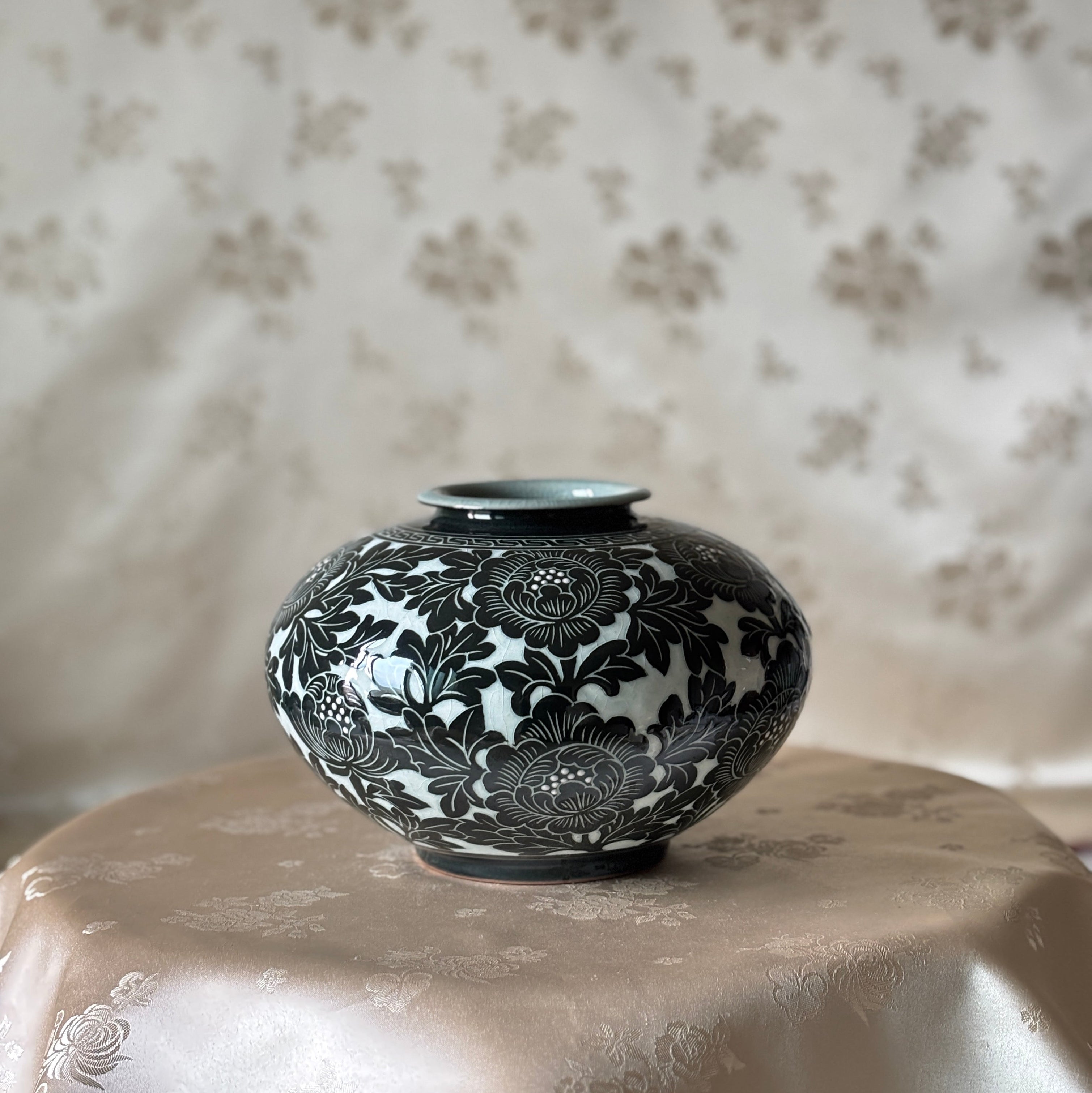 A small round ceramic vase with a black floral pattern on a white glaze, displayed on a draped beige satin cloth against a matching patterned fabric backdrop.