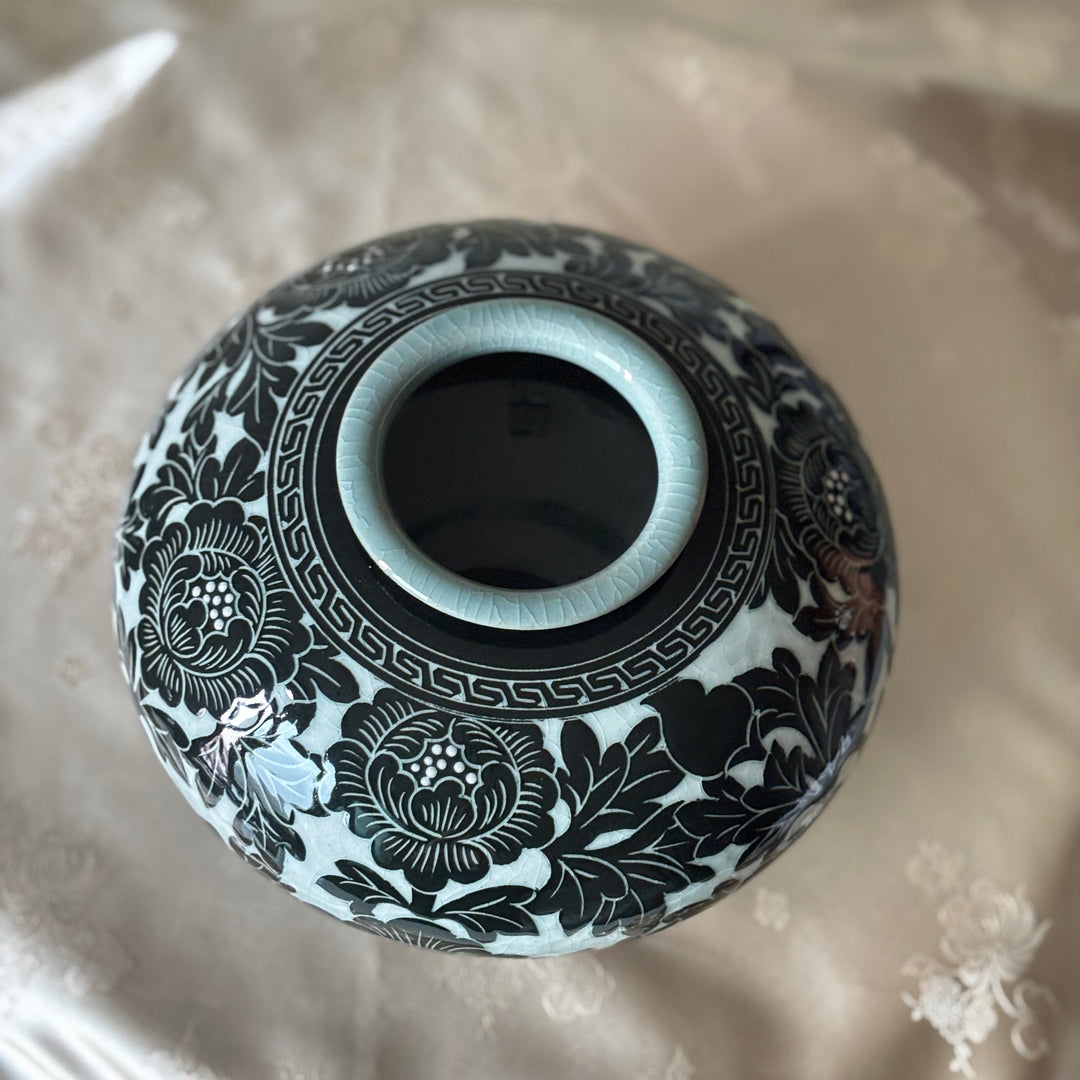 A round ceramic vase with a black floral and geometric pattern on a white glaze, photographed from above on a beige satin fabric background.