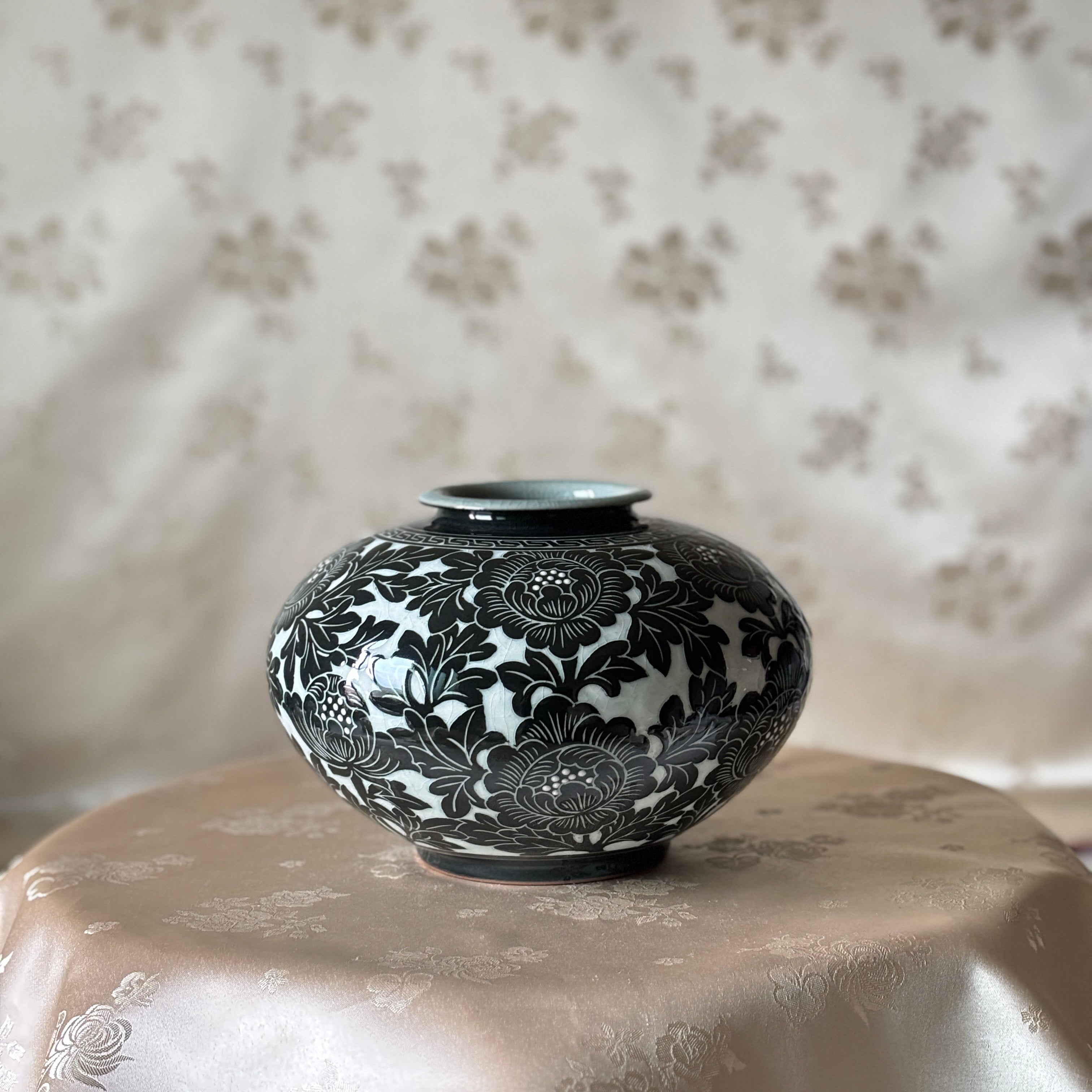A small round ceramic vase with a glossy black floral pattern on a white glaze, displayed on a draped beige satin cloth against a matching patterned fabric backdrop.