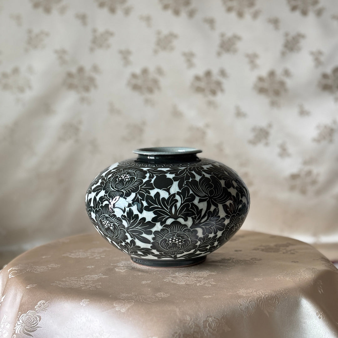 A small round ceramic vase with a glossy black and white floral pattern, displayed on a draped beige satin surface against a matching patterned fabric backdrop.