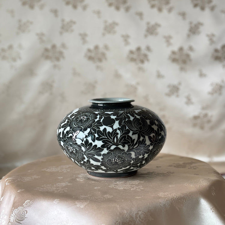 A small round ceramic vase with a glossy black and white floral pattern, displayed on a draped beige satin surface against a matching patterned fabric backdrop.