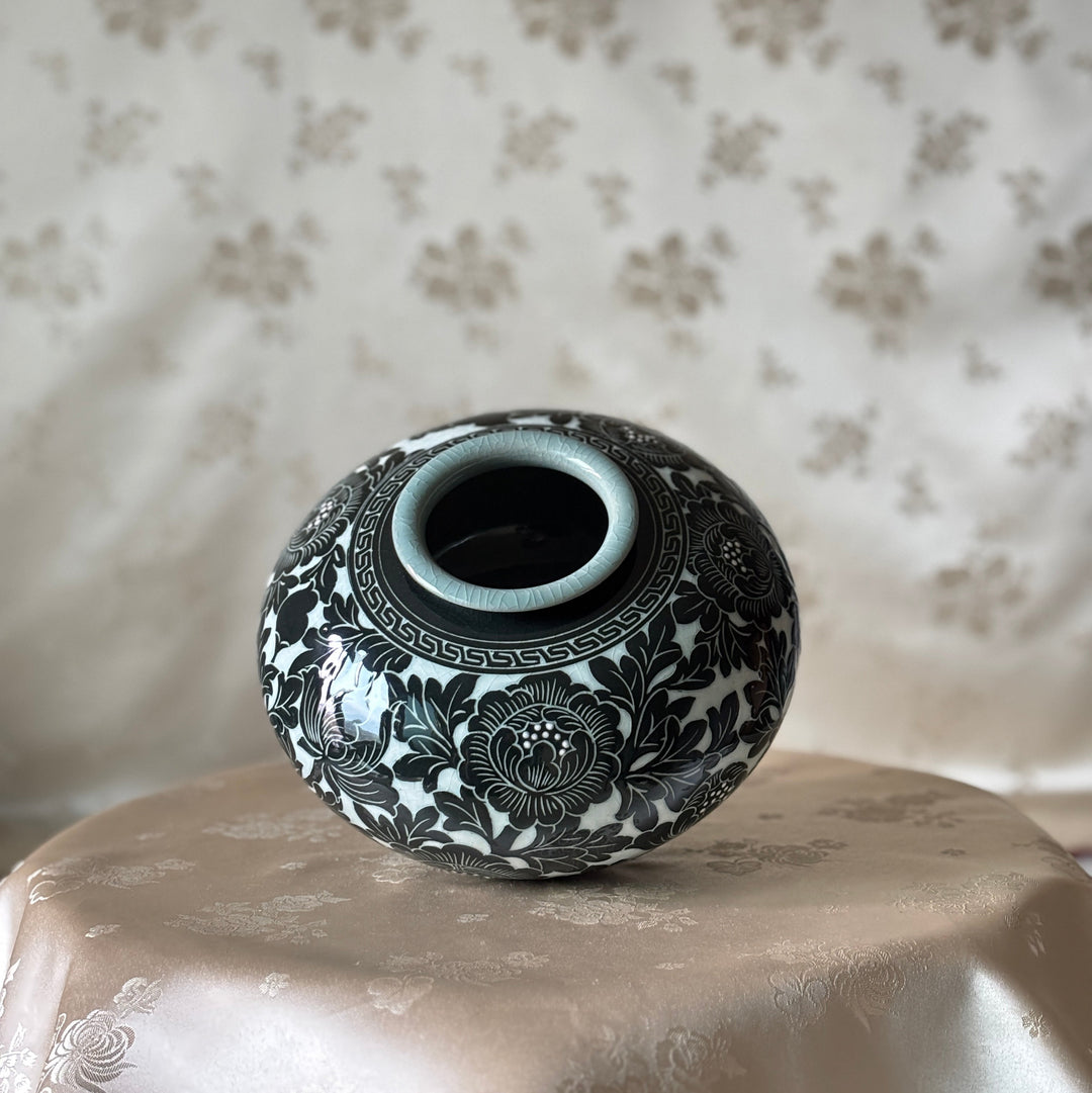 A small round ceramic vase with a black floral pattern on a white glaze, displayed on a draped beige satin cloth against a matching patterned fabric backdrop.