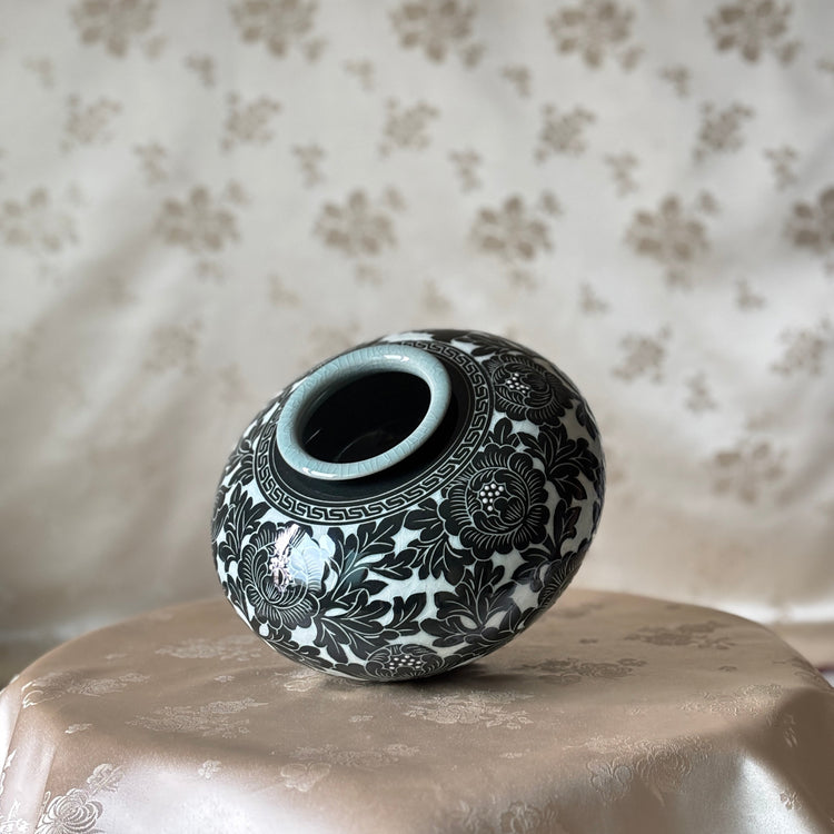 A small decorative ceramic vase with a black floral pattern on a white glaze, displayed on a draped beige satin cloth against a matching patterned fabric backdrop.