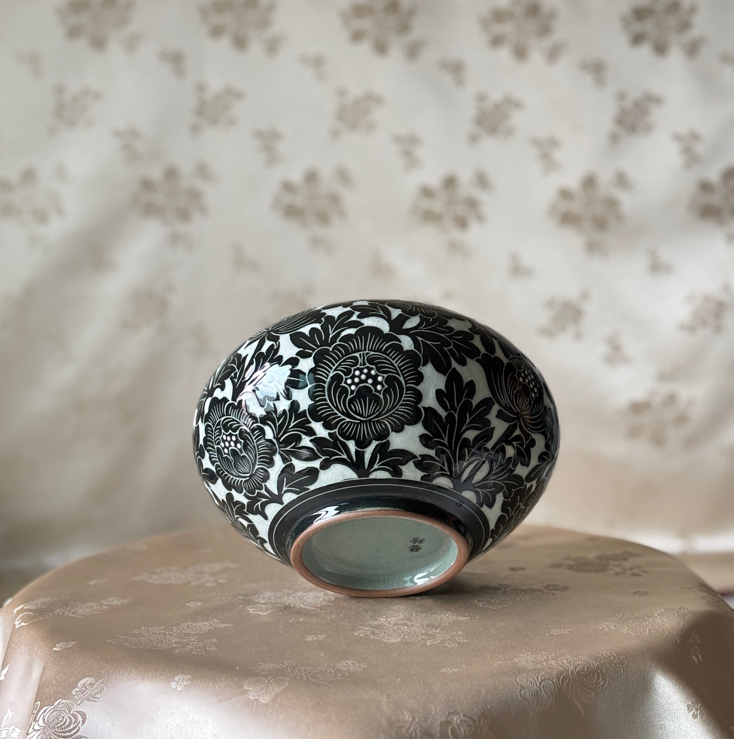 A small round ceramic bowl with a black floral pattern on a celadon glaze, displayed upside down on a beige satin-covered surface against a patterned beige fabric backdrop.