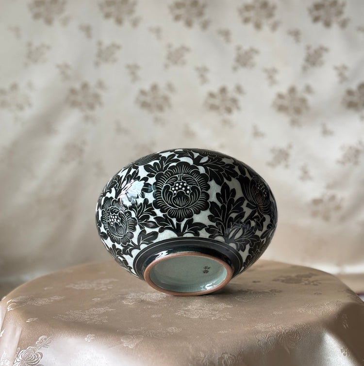 A small round ceramic bowl with a black floral pattern on a celadon glaze, displayed upside down on a beige satin-covered surface against a patterned beige fabric backdrop.