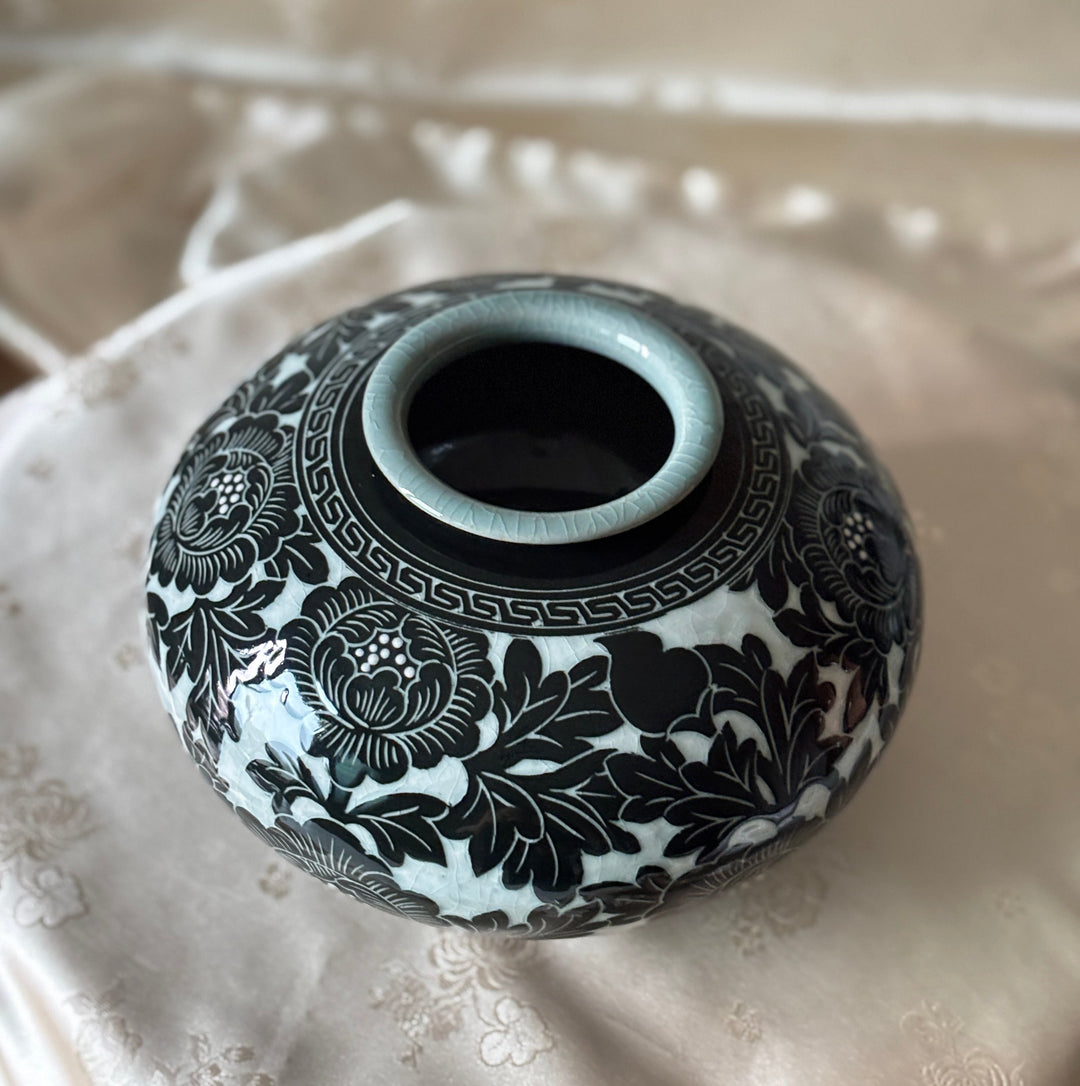 A small round ceramic vase with a glossy black floral pattern on a white glaze, featuring a pale blue-green rim, displayed on a beige embroidered fabric surface.