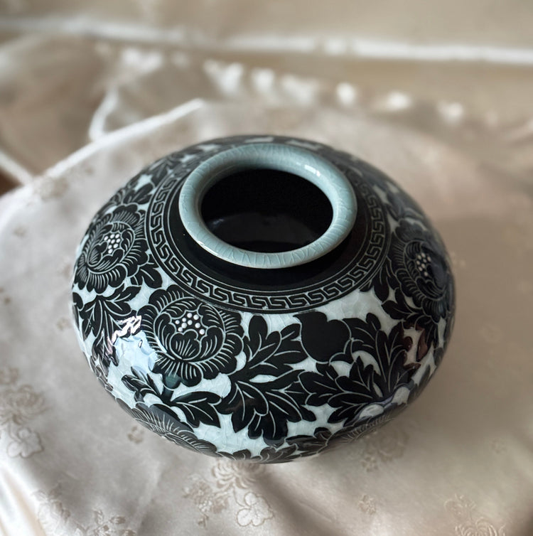 A small round ceramic vase with a glossy black floral pattern on a white glaze, featuring a pale blue-green rim, displayed on a beige embroidered fabric surface.