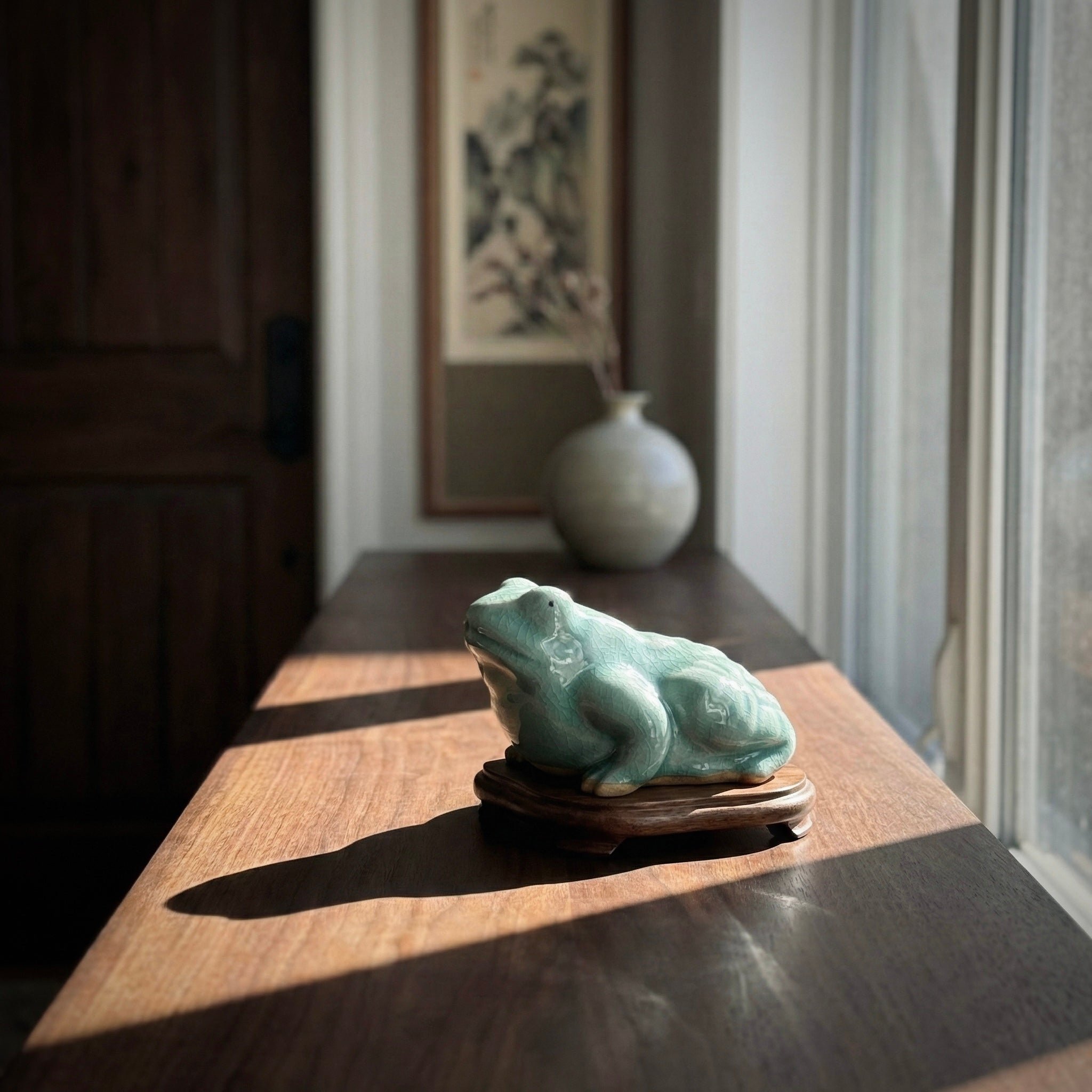 Green frog sculpture on a wooden stand in a room with a vase and framed picture.