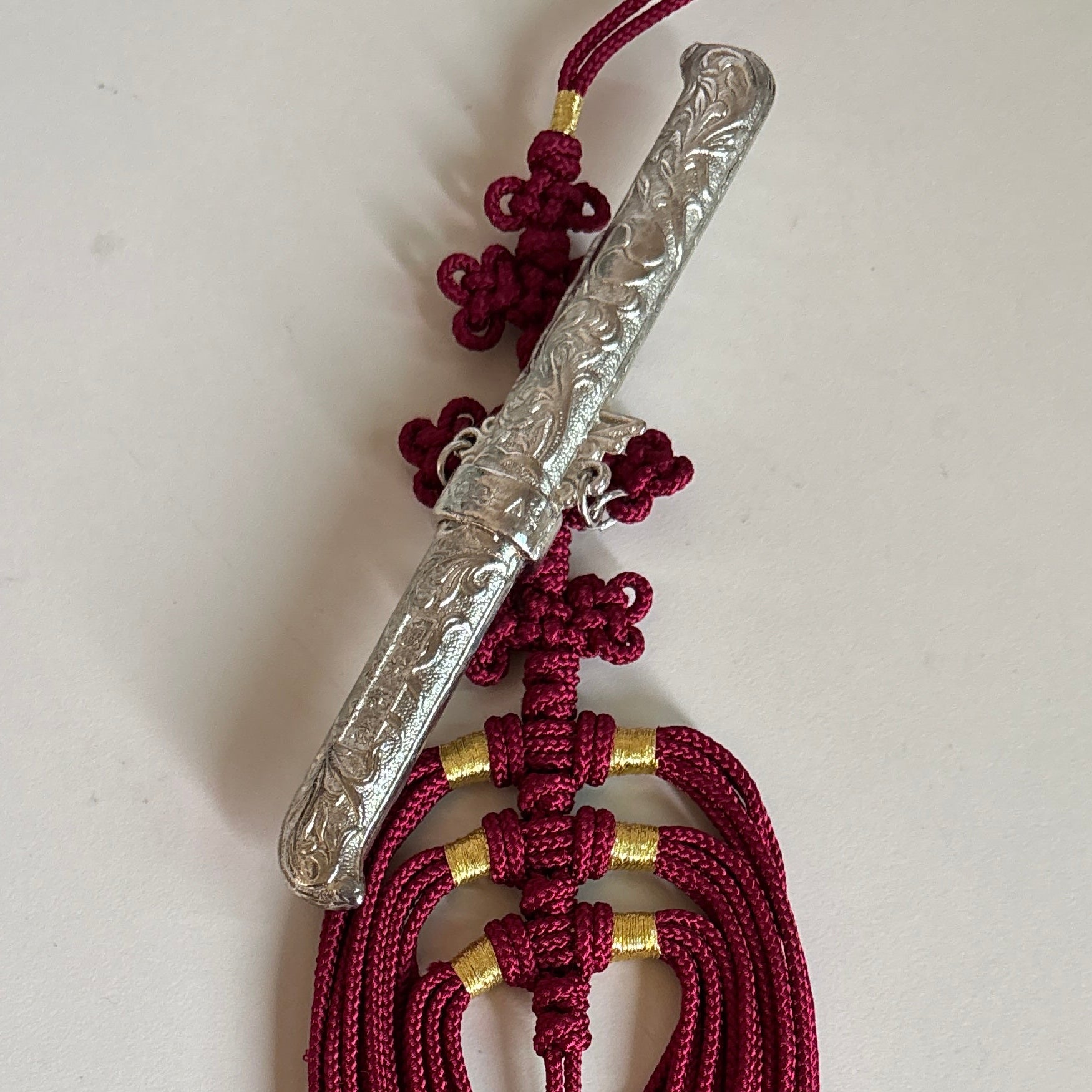 A traditional Korean tassel ornament featuring a carved silver-colored metal rod, deep red braided cord, and small gold spacer rings, displayed on a plain off-white surface.