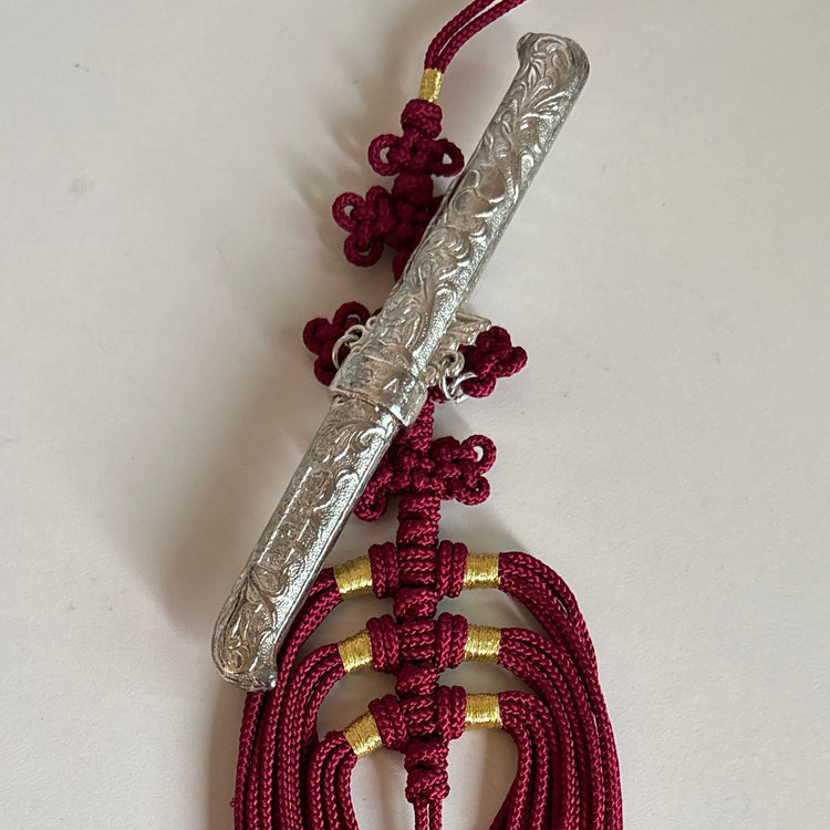 A traditional Korean tassel ornament featuring a carved silver-colored metal rod, deep red braided cord, and small gold spacer rings, displayed on a plain off-white surface.