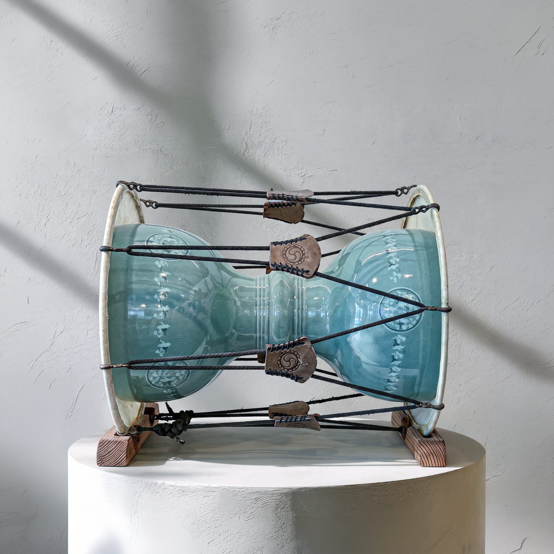 A handmade ceramic drum with a glossy light blue-green glazed body, mounted on a white cylindrical pedestal against a textured light gray wall with soft diagonal shadows.