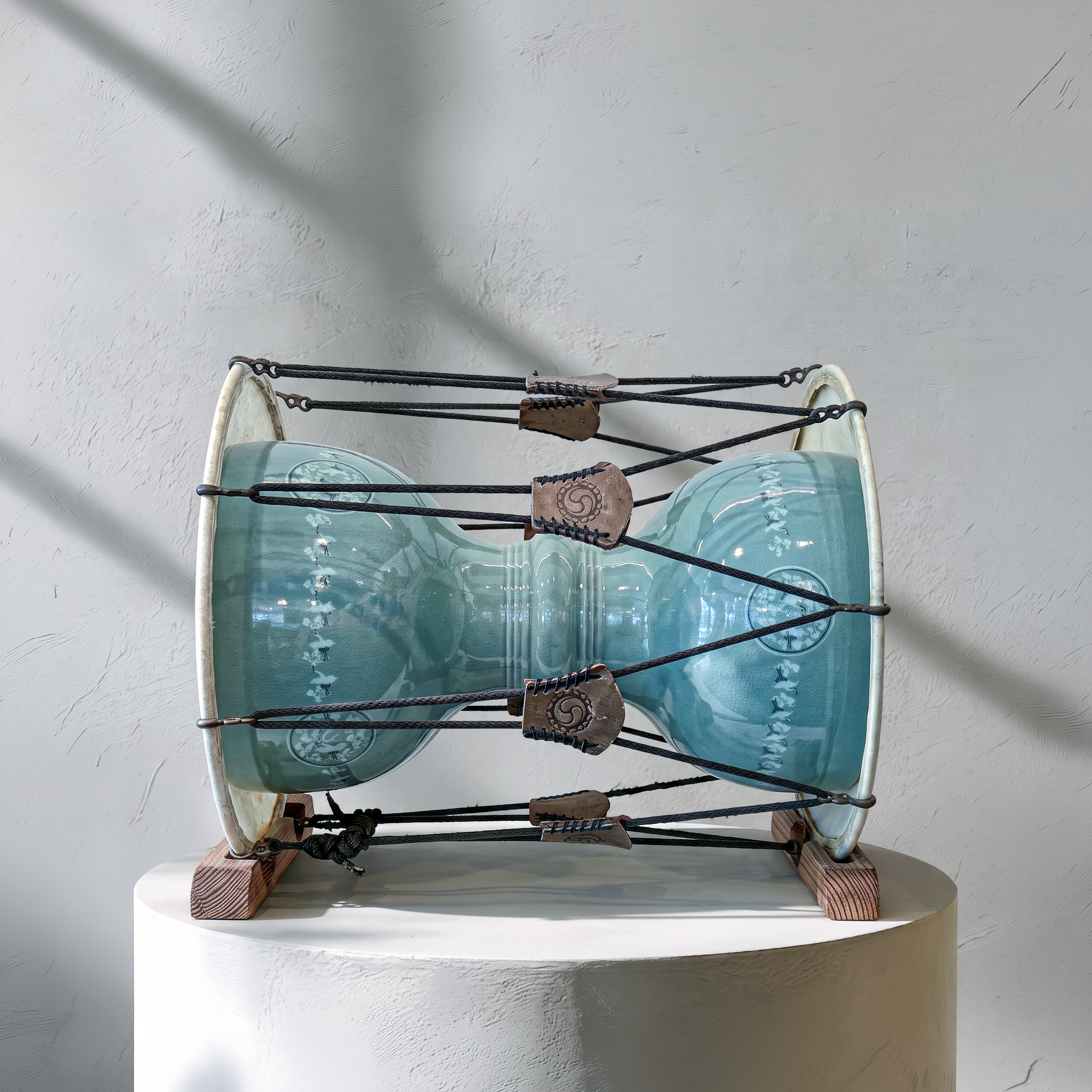 A handmade ceramic drum with a glossy light blue-green glazed body, mounted on a white cylindrical pedestal against a textured light gray wall with soft diagonal shadows.