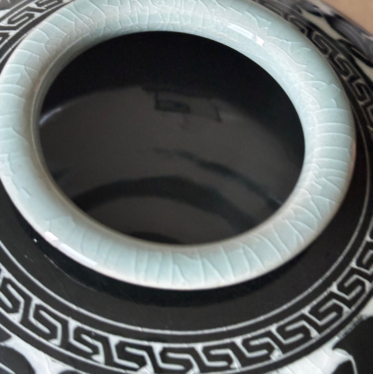 Close-up of a ceramic vase or planter with a glossy black exterior featuring a white geometric border pattern, a pale celadon-green glazed rim, and a dark interior.