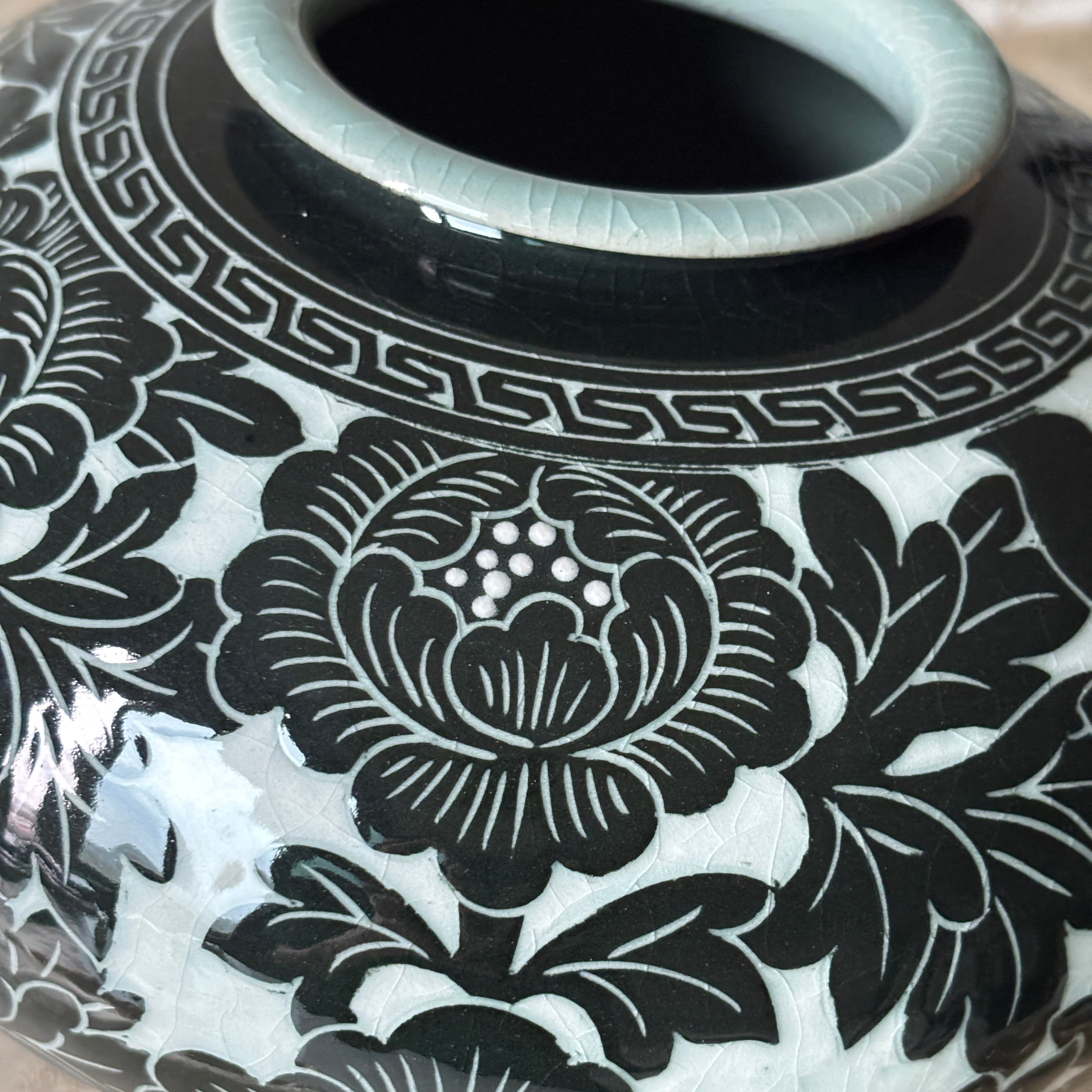 Close-up of a glossy ceramic vase with a black floral pattern on a white glaze, featuring a pale blue-green rim and a peony motif 