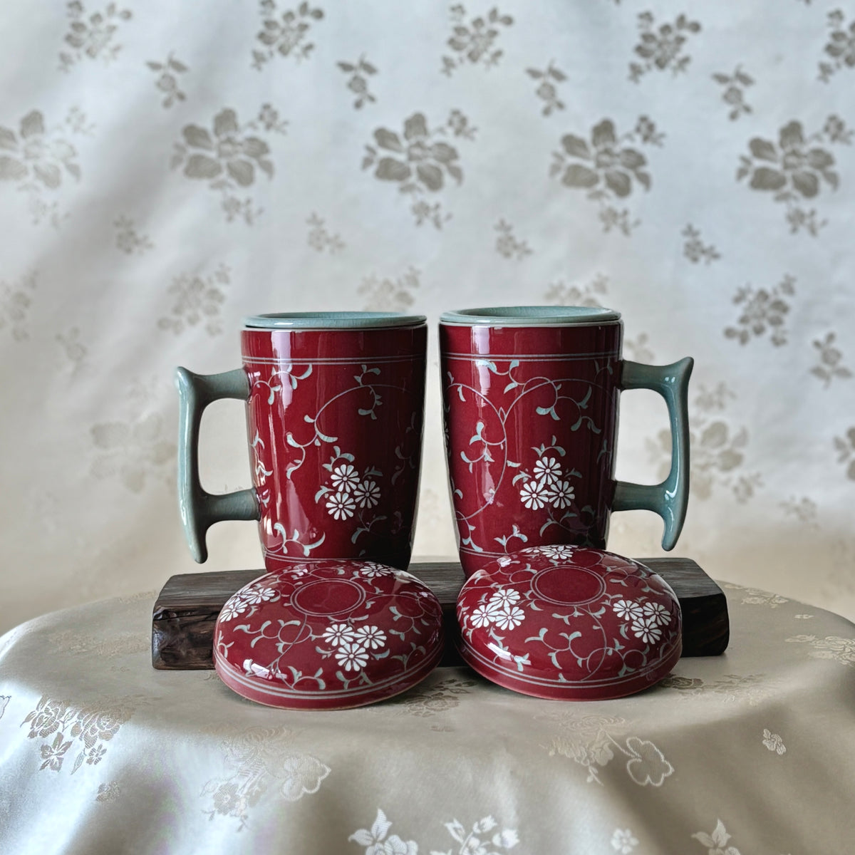 Two red celadon mugs with inlaid chrysanthemum and vine motifs, placed on a surface with a floral pattern in the background.