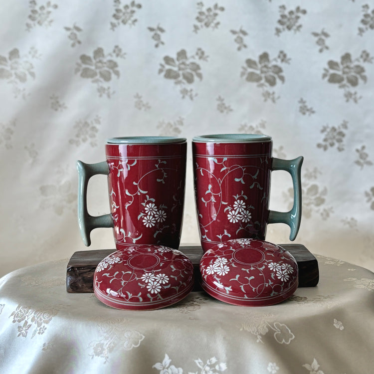 Two red celadon mugs with inlaid chrysanthemum and vine motifs, placed on a surface with a floral pattern in the background.