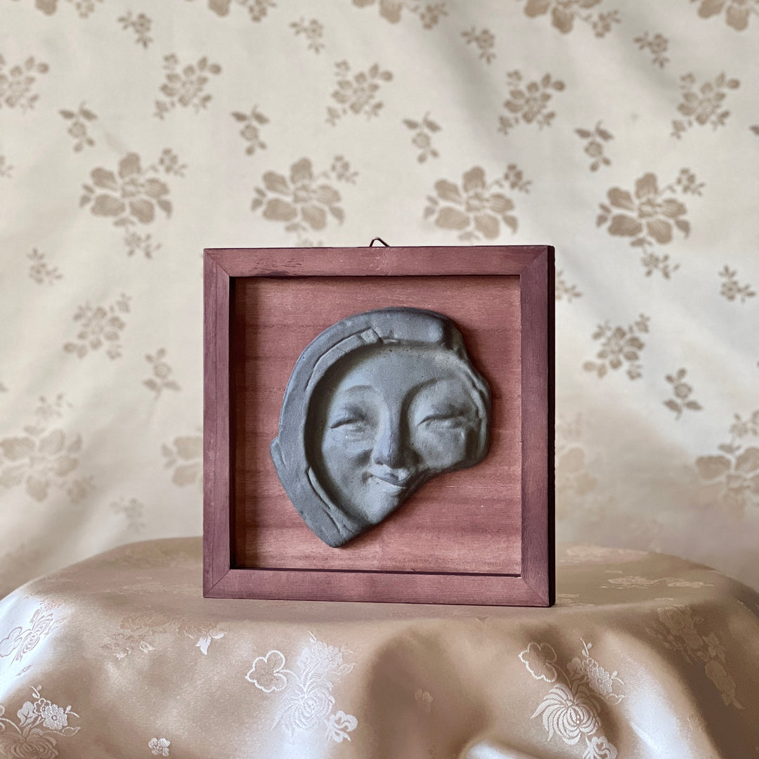 Decorative wall art with a face tile in a wooden frame on a floral-patterned surface.