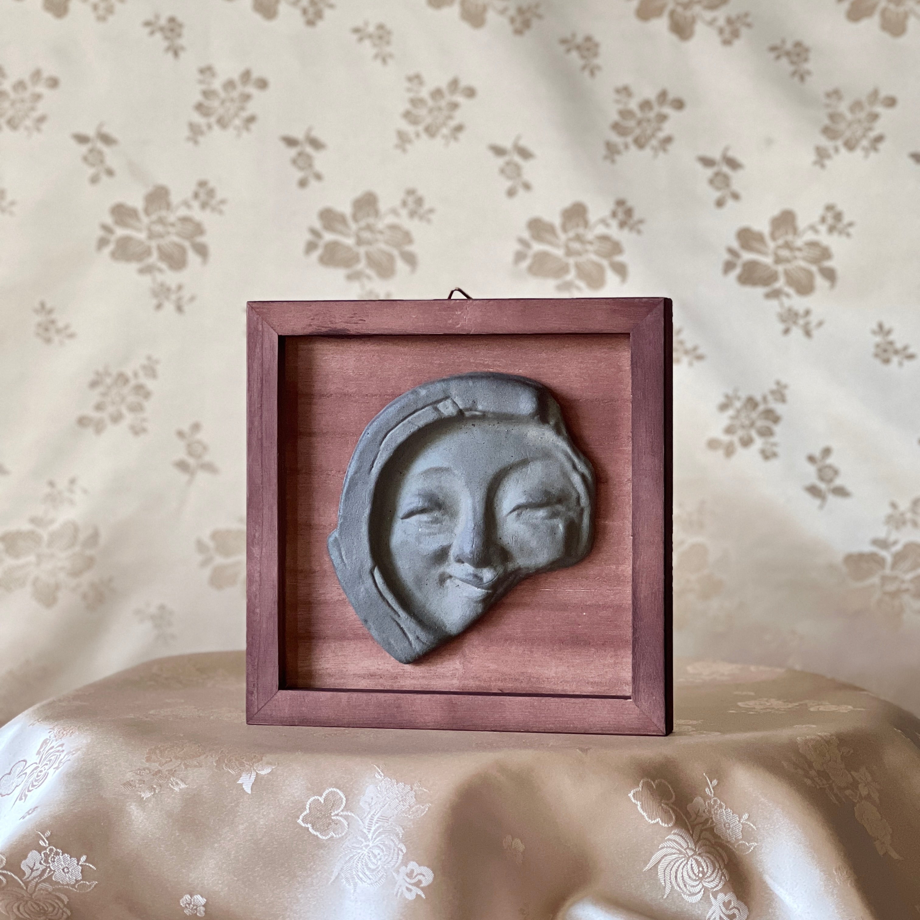 Decorative wall art with a face tile in a wooden frame on a floral-patterned surface.