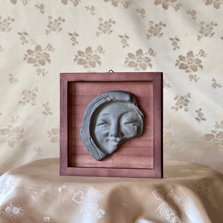 Decorative wall art with a face tile in a wooden frame on a floral-patterned surface.