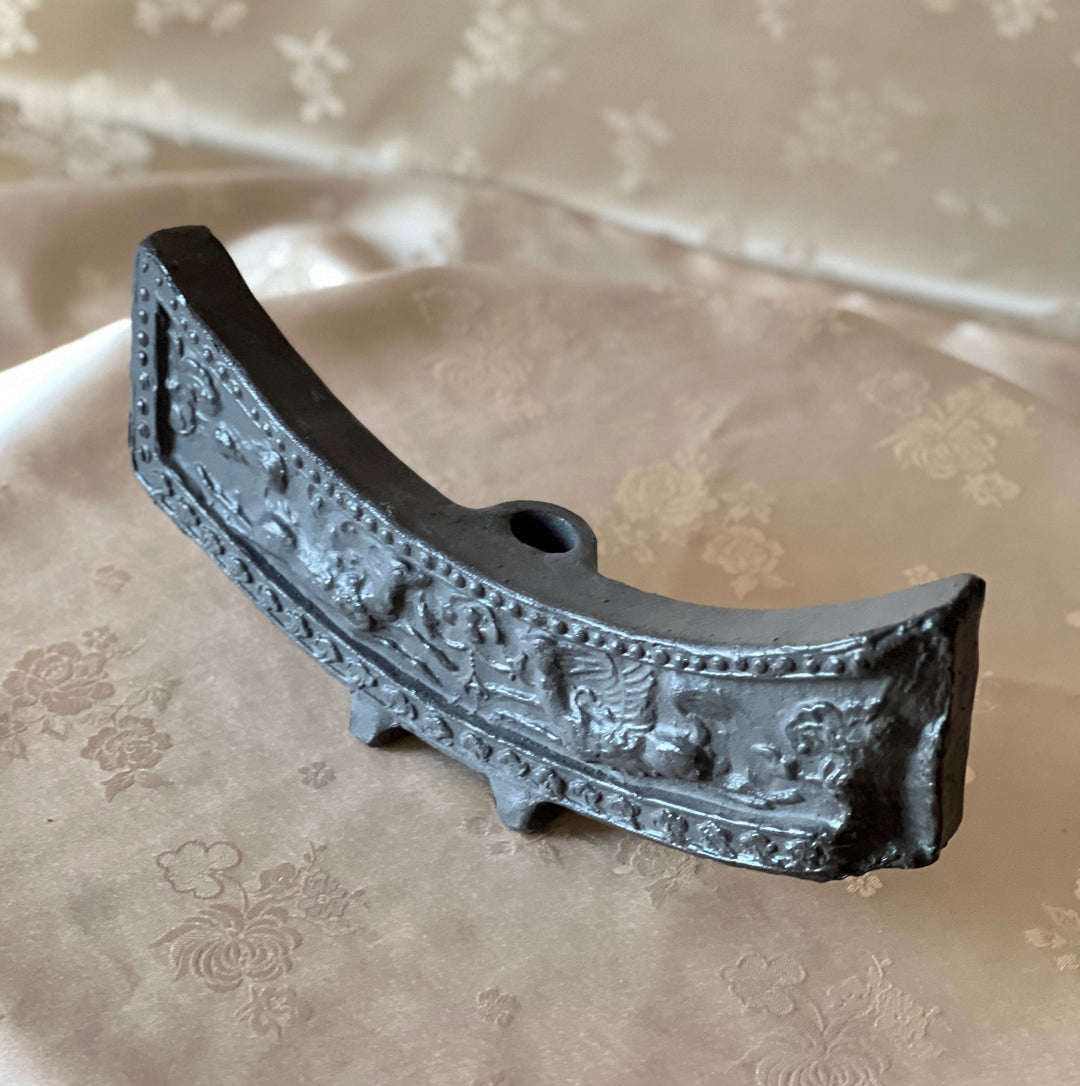 An antique curved metal object with intricate embossed relief patterns, displayed on a beige patterned fabric surface.