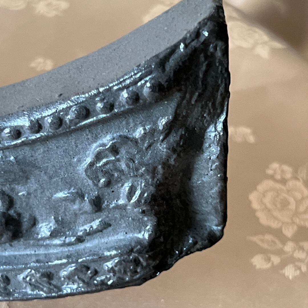 A close-up of a dark gray, textured decorative molding piece or architectural trim sample, showing embossed floral and beaded details, set against a beige patterned fabric background.