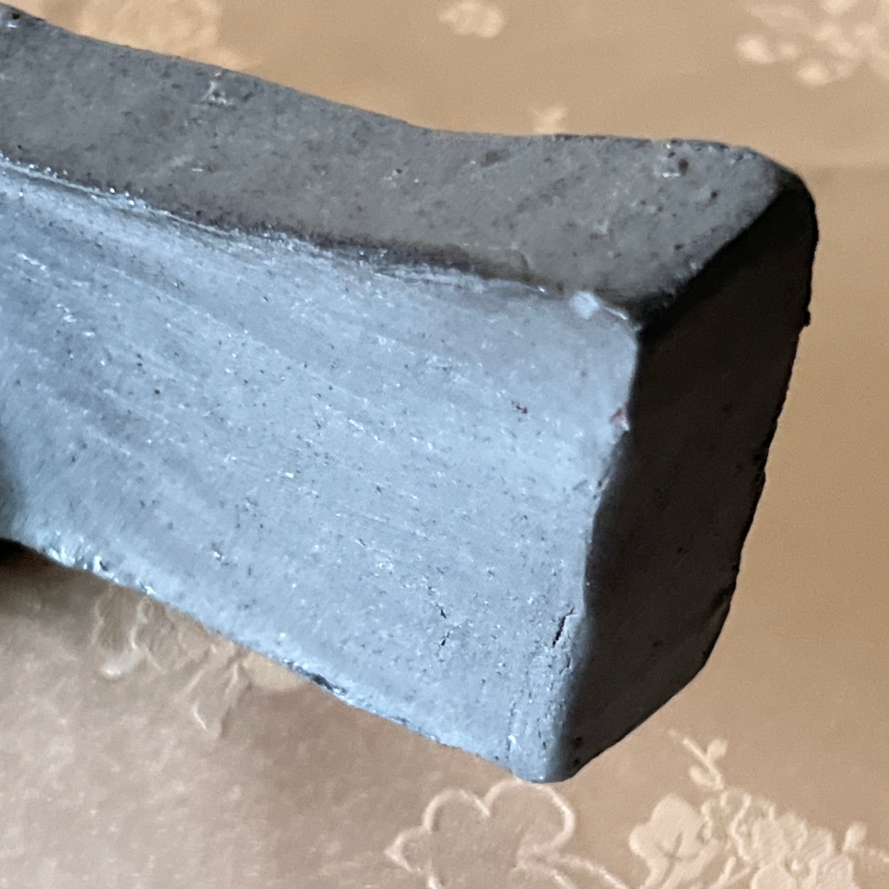 A close-up of a dark gray rectangular block or tile with a rough, matte texture, resting on a beige patterned fabric surface.