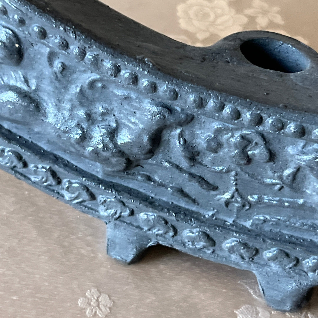 Close-up of a dark gray cast iron pipe bend with embossed floral and scrollwork patterns, resting on a beige patterned fabric surface.