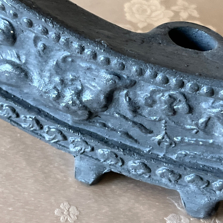 Close-up of a dark gray cast iron pipe bend with embossed floral and scrollwork patterns, resting on a beige patterned fabric surface.