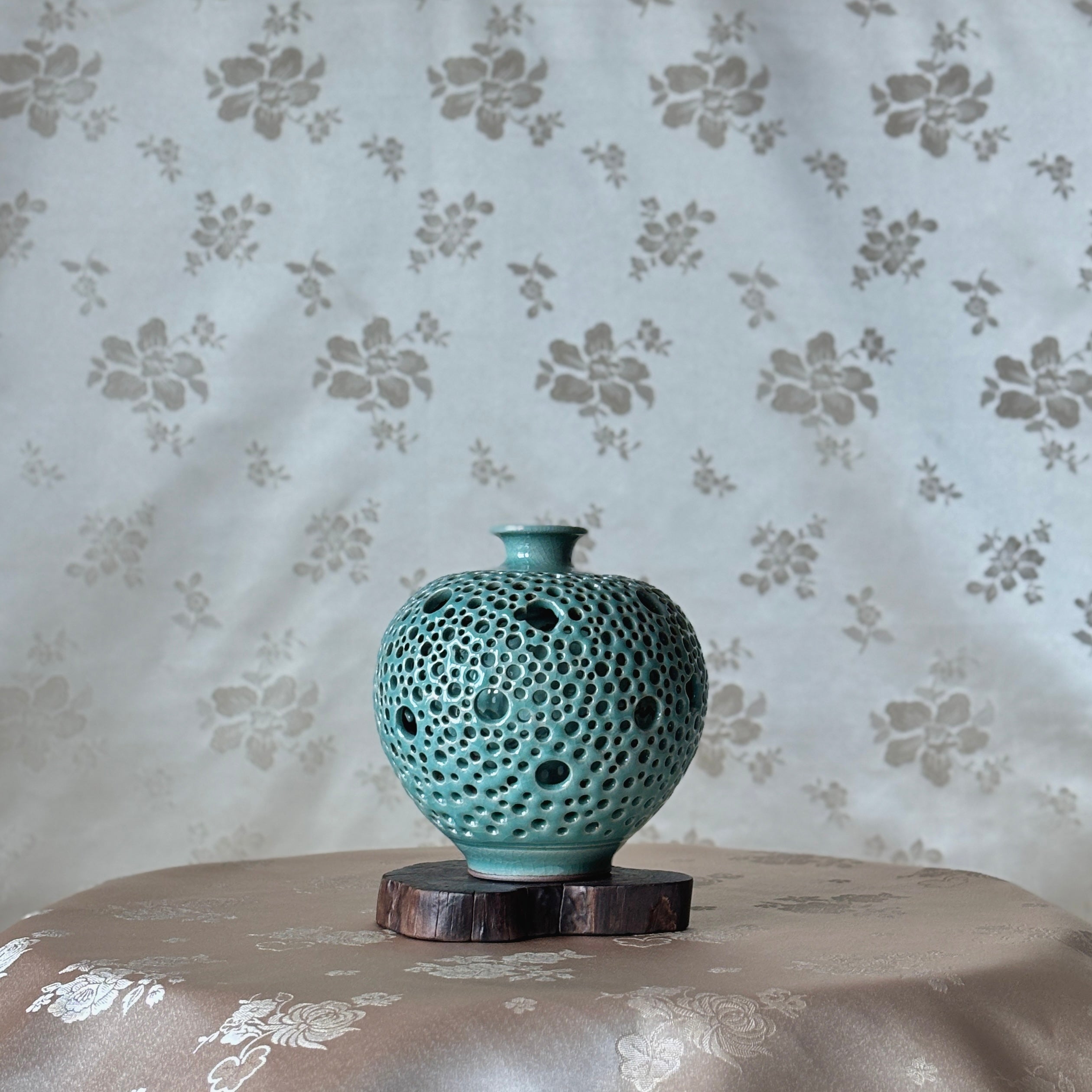 jade ceramic vase with textured surface on a wooden stand against a floral-patterned wall.